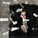 Nevető napszemüveges gyerek fekete fotelben, bankjegy eső, öröklési jog